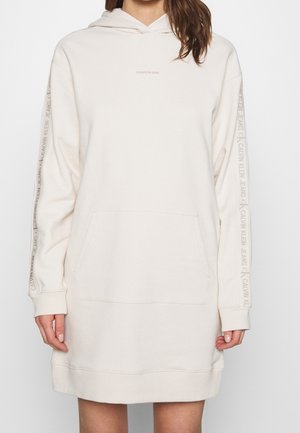 Femme portant une robe à capuche crème Calvin Klein avec poche kangourou et texte de la marque sur les manches et la poitrine.