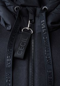 Veste matelassée noire avec fermeture zip, présentant un tissu texturé, une capuche lisse contrastante et des sangles tissées avec des noms de marque en relief.