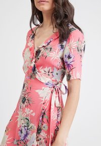 Robe portefeuille à fleurs roses en tissu léger, avec manches courtes, décolleté en V, détails à volants et ceinture nouée à la taille.