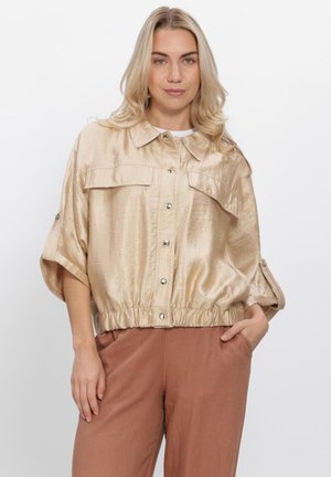 Blondine vrouw met een glanzende beige jas met knopen en elastische taille, en roestkleurige broek, staand met één hand in de zak.
