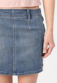 Jupe mini en denim bleu clair, présentant une forme ajustée, une fermeture éclair à l'avant et de subtils détails de couture. Texture lisse, sans motifs visibles.