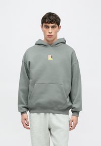 Γκρι φλις hoodie με τσέπη καγκουρό και κουκούλα. Διαθέτει μικρό πολύχρωμο λογότυπο στο στήθος. Το μοντέλο φοράει ανοιχτό γκρι φόρμα.