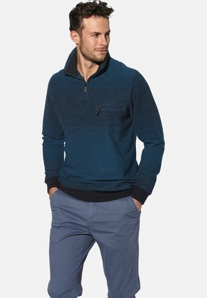 Mann mit kurzen, lockigen Haaren, der einen dunkelblauen Pulli mit Viertelreißverschluss und hellblauen Hosen trägt, die Hände in den Taschen, nach rechts schauend.