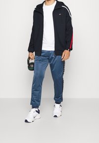 Schwarze Zip-Hoodie mit rotem Akzent; weißes T-Shirt; blaue Satin-Sporthose mit seitlichem Branding; weiße und marineblaue Sportschuhe. Hält eine Kettlebell.