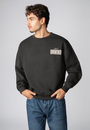 Schwarzer Pullover aus Baumwolle mit einem Rundhalsausschnitt, auf der Brust mit einer beige Grafik "Zeit zum Entspannen" versehen. Kombiniert mit blauen Jeans.