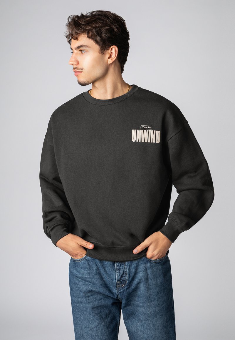 Schwarzer Pullover aus Baumwolle mit einem Rundhalsausschnitt, auf der Brust mit einer beige Grafik "Zeit zum Entspannen" versehen. Kombiniert mit blauen Jeans.