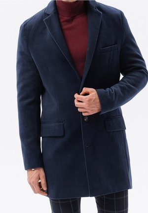 Homme portant un manteau en laine bleu marine sur un pull à col roulé bordeaux, boutonnant le manteau, debout sur fond blanc.