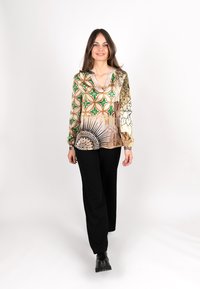 Musterbluse mit grünen und goldenen geometrischen Designs, V-Ausschnitt, langen Ärmeln. Kombiniert mit schwarzen weiter geschnittenen Hosen und klobigen schwarzen Stiefeln.