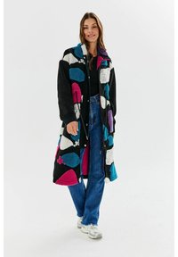 Czarny długi płaszcz z kolorowymi geometrycznymi wzorami w odcieniach różu, niebieskiego i białego. Miękka, teksturowana tkanina z zamkiem błyskawicznym z przodu i kieszeniami.
