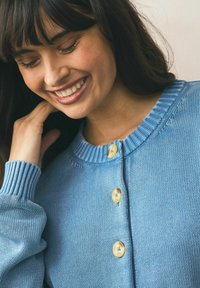 Cardigan tricotat de culoare bleu deschis, cu guler ribbit, mâneci lungi și trei nasturi mari de culoare crem pe față. Detalii texturate ale materialului sunt vizibile.