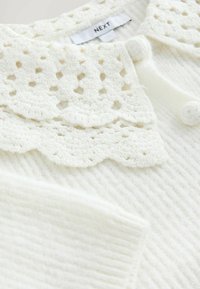 Hvid pullover med ribbet tekstur. Har en dekorativ blondekrave med udskårne mønstre og knapdetaljer. Blødt stof.