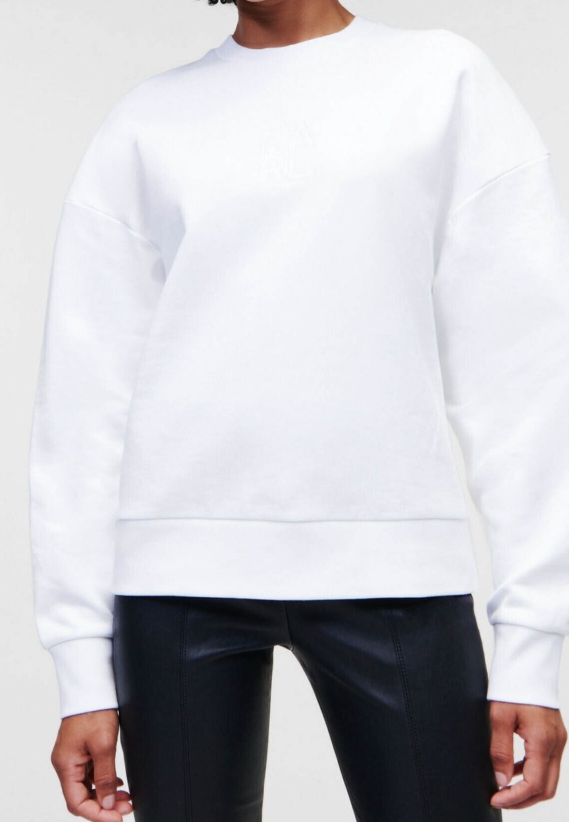 Personne portant un sweat-shirt blanc uni à manches longues et un pantalon en cuir noir, recadré au niveau du cou et des cuisses.