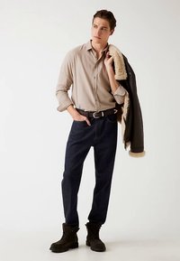 Chemise beige clair boutonnée, pantalon en jean bleu marine et veste noire doublée en peau lainée posée sur l'épaule. Bottines marron à lacets complètent la tenue.