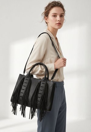 Junge Frau in beiger Bluse und blauer Hose trägt eine schwarze Lederhandtasche mit langen Fransen am Schulterriemen.