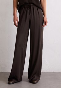 Wijdvallende broek in donkerbruin, gemaakt van een zachte, lichte stof met een elastische tailleband en verstelbaar trekkoord. Minimale details.