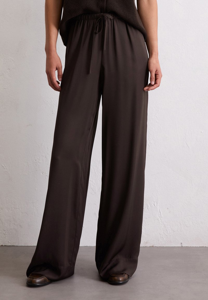 Wijdvallende broek in donkerbruin, gemaakt van een zachte, lichte stof met een elastische tailleband en verstelbaar trekkoord. Minimale details.