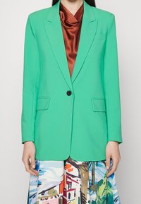 Blazer verde realizzato in tessuto liscio, con revers a punta, tasche frontali e chiusura a singolo bottone nero, abbinato a una gonna colorata.