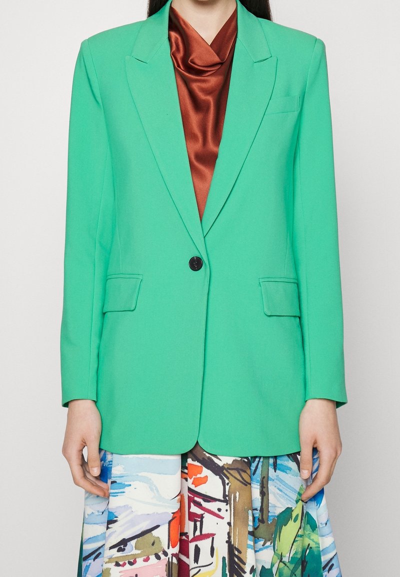 Blazer verde realizzato in tessuto liscio, con revers a punta, tasche frontali e chiusura a singolo bottone nero, abbinato a una gonna colorata.