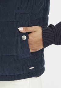 Mörkblå kordflersväst med en texturerad yta, med en silverknapp och en liten metallisk logotagg på fållen.