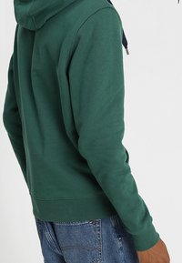 Green hooded sweatshirt with a textured finish, featuring a drawstring hood, ribbed cuffs, and hem. Worn with blue denim jeans.