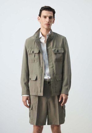 Hombre con chaqueta verde oliva claro y pantalones cortos cargo a juego, con camisa blanca, sobre un fondo claro y liso.