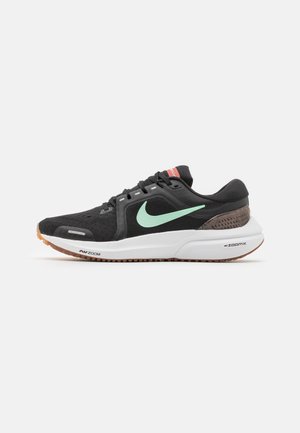 Schwarz-weißer Nike-Laufschuh mit grünem Swoosh, schwarzen Schnürsenkeln, gepolsterter Sohle und braunem Profil, seitlich auf weißem Hintergrund abgebildet.