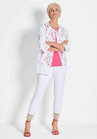Weiße Spitzenjacke mit floralen Ausschnitten über einem rosafarbenen Oberteil, kombiniert mit weißen Hosen mit dekorativem Saum und rosa Riemchensandalen.