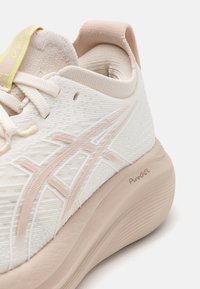 Zapatilla de running blanca y rosa claro con parte superior de tejido texturizado, acentos blancos, cuello acolchado y la marca "PureGEL" en la suela.