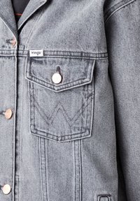 Grå denimjacka med knäppning, två bröstfickor med dekorativ sömnad och en liten logotags framtill.
