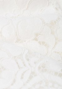 Bílá krajková látka s texturovaným vzorem abstraktních tvarů, s hladkým, jemným povrchem a složitými stehy.