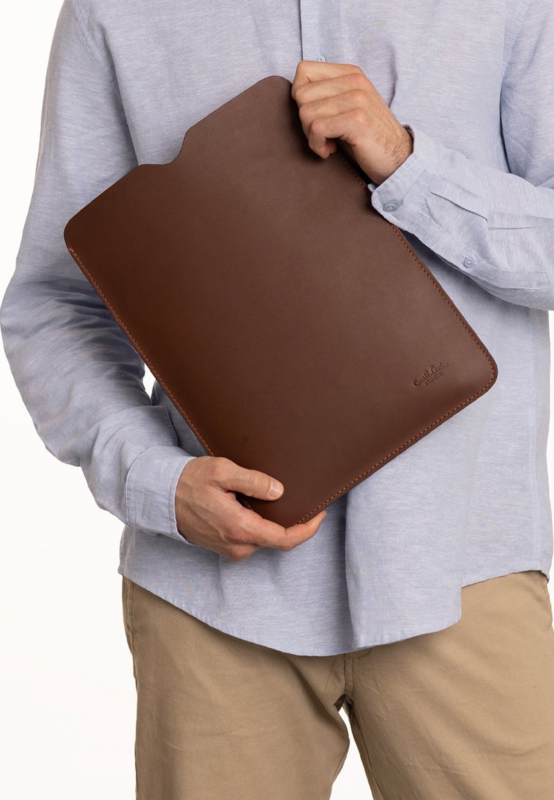 Brązowy skórzany holder na dokumenty o gładkim wykończeniu, lekko zaokrąglonych krawędziach i wycięciu dla łatwego dostępu, trzymany przez osobę w jasnej koszuli.