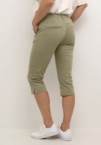 Zelene kapri hlače s slim fit krojem, z dvema zadnjima žepoma in stranskima razporkoma na robu. Nosi se z belimi supergami.