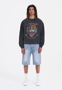 Felpa nera con una grafica di una tigre colorata e testo sulla parte anteriore, abbinata a pantaloni shorts in denim chiaro e sneakers bianche.