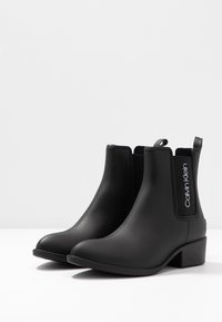 Bottines Chelsea noires en caoutchouc avec une texture lisse, comportant des panneaux latéraux élastiques et un talon épais. Logo Calvin Klein sur le côté.