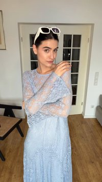 Frau in einem hellblauen Spitzenkleid und mit weißen Sonnenbrillen auf dem Kopf, die mit verschränkten Armen in der Nähe einer Glastür und eines Holzbodens steht.