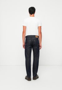 Mann, der dunkelblaue Jeans und ein weißes T-Shirt trägt, steht mit dem Rücken vor einem schlichten weißen Hintergrund.