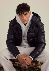 Veste matelassée noire avec capuche et fermeture éclair, superposée à un sweat-shirt gris. Modèle assis, portant un pantalon de survêtement gris, mains jointes.