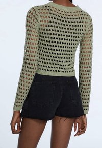 Maglione verde a maglia aperta con maniche lunghe, caratterizzato da un collo rotondo e una vita aderente, abbinato a pantaloni corti di jeans neri, che mostrano dettagli delle tasche.