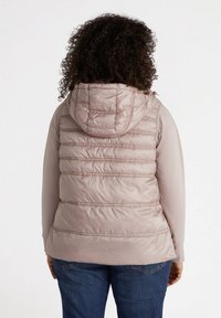 Vaaleanpunainen puffi liivi, jossa on huppu, vaakasaumat, istuva leikkaus ja sileä pinta. Päällä pitkähihaisen paidan kanssa.