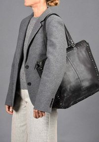 Zwarte leren tote bag met een gestructureerde afwerking, met zilveren studs langs de randen, gedragen door een persoon in een grijze blazer en sweater.