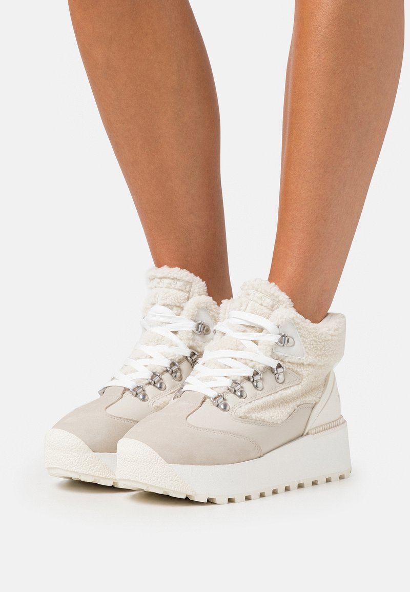 Baskets blanches montantes avec semelles épaisses, œillets métalliques et doublure en polaire douce portées aux pieds d'une personne.