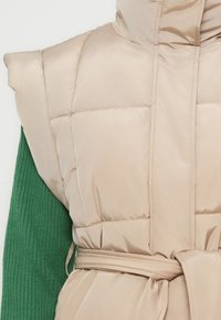 Bežna podložena brezrokavnik s pasom, nošena čez zeleni rebrasti pulover z dolgimi rokavi na beli podlagi.