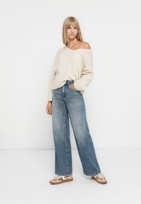 Beige oversized Pullover mit V-Ausschnitt und gerippten Bündchen, kombiniert mit hoch sitzenden, hellblauen weitem Jeans und gemusterten Sneakers.