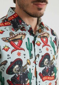 Chemise à manches courtes avec un fond bleu clair ornée d'imprimés colorés sur le thème mexicain, y compris des crânes, des sombreros, des cactus et des piments.