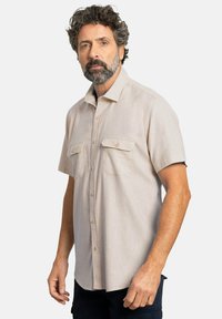 Camisa de manga corta color beige claro con una textura sutil, dos bolsillos en el pecho y botones en tono marrón. Bajo recto y diseño de cuello clásico.