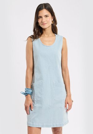 Femme portant une robe sans manches à rayures bleu clair et blanc avec des poches frontales, des boucles d'oreilles créoles dorées, et un foulard bleu noué autour du poignet.