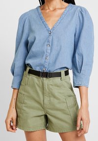 Camicia di denim azzurro chiaro con scollatura a V e apertura con bottoni, abbinata a pantaloni corti verde oliva con tasche laterali. Cintura nera che accentua l'outfit.