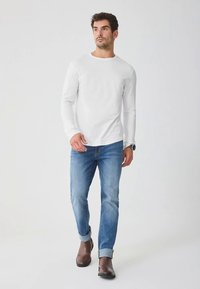 Biela košeľa s dlhými rukávmi, modré rifľové nohavice, hnedé kožené topánky. Model stojí s neutrálnym výrazom na jednoduchom pozadí.