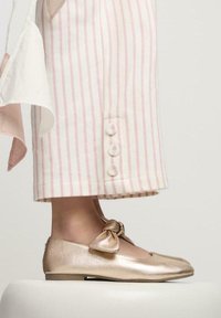 Ballerine metalliche dorate con un dettaglio a fiocco annodato, abbinate a pantaloni larghi a righe rosa e bianche con dettagli in bottoni.