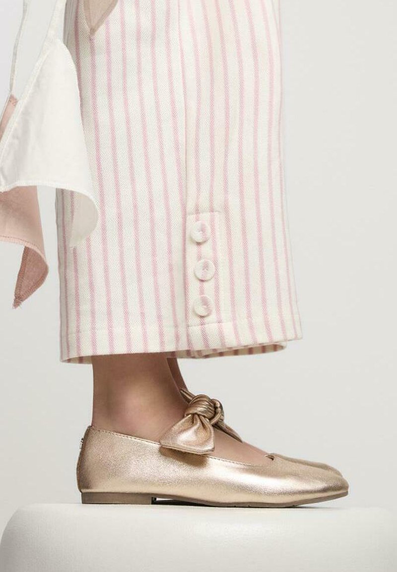 Ballerine metalliche dorate con un dettaglio a fiocco annodato, abbinate a pantaloni larghi a righe rosa e bianche con dettagli in bottoni.